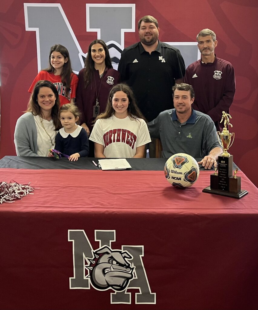 New Albany Lady Bulldog Soccer Player Caroline King (front center) is surrounded by family and coaches as she signs to play soccer for Northwest Mississippi Community College on Thursday, February 17. Pictured front row l-r:  Bailey King, Elizabeth King, Caroline King, Geoffrey KingBack row l-r:  Sara Jane King; Katelyn Robbins, NAHS Assistant Soccer Coach; Cody Stubblefield, NAHS Athletic Director; Bert Anderson, NAHS Head Girls Soccer Coach.
